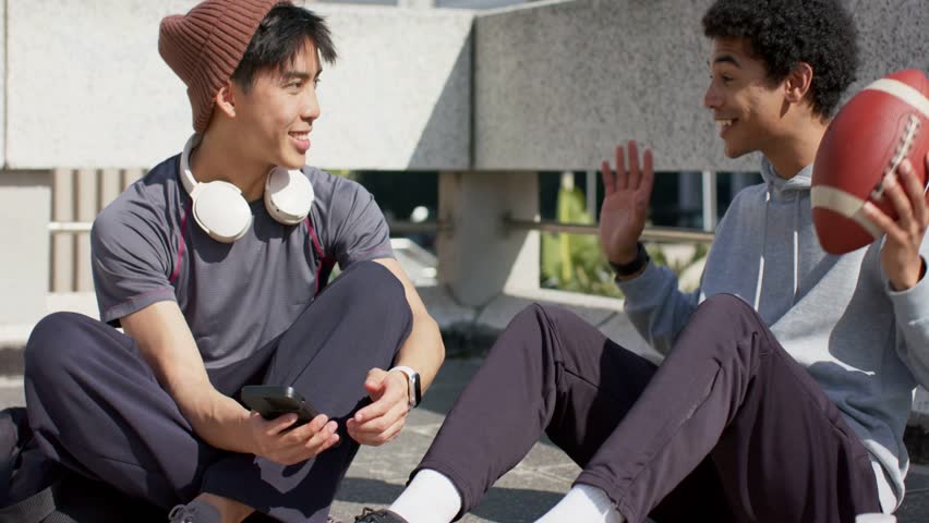 Two men on ledge, left man with smartphone, prompt appearing and expanding chatbot tech explanation. Duo, friends, outdoor, urban, plaza, daylight, conversation
