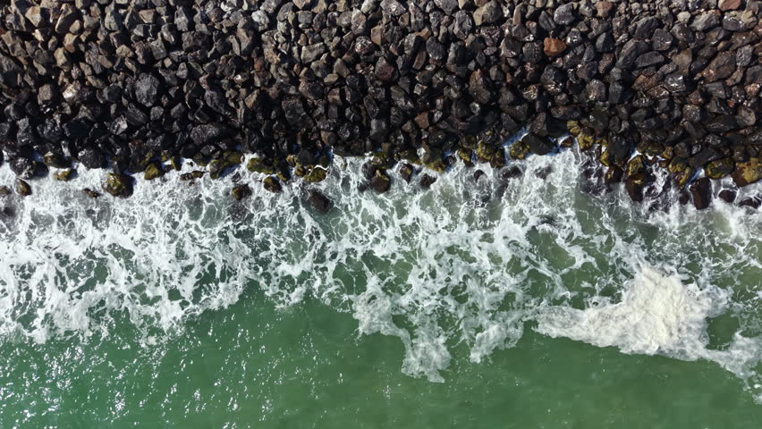 Waves flow over rocks at the edge of a beach area in a coastal setting. The scene captures the interaction between water and stones during daytime with sunlight reflecting on the surface.