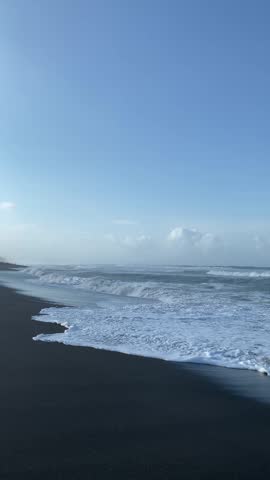 Beautiful scenery of a black sand beach with white ocean waves under a clear blue sky. A serene tropical coastal landscape during the day.