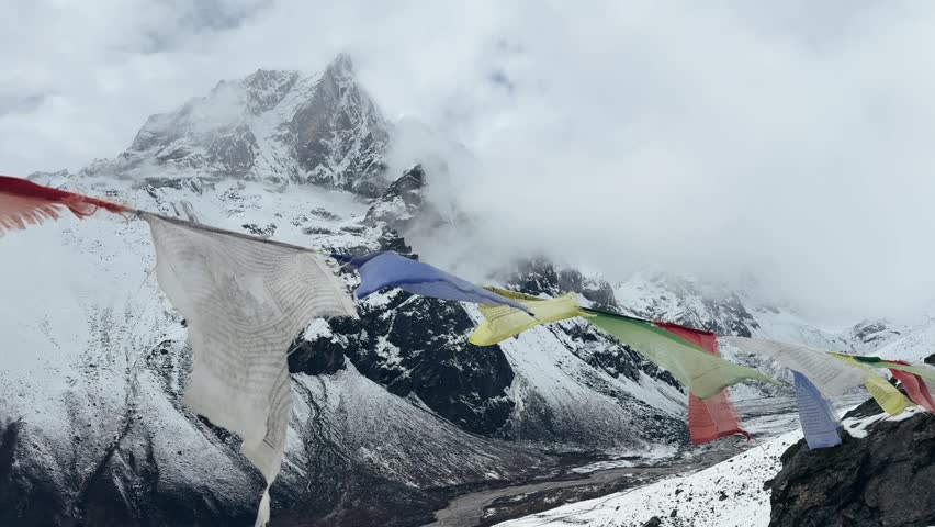 Tibetan prayer flags fluttering against cloudy snowy mountains in Everest region, Nepal. Dramatic Himalayan landscape with mist and moving clouds, atmospheric slow motion shot