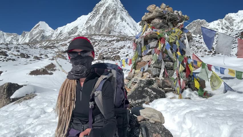 Female trekker with backpack, dreadlocks and face covered records selfie video near memorial on Everest Base Camp trail, snowy Himalayan mountains, high altitude around 5000m, Lobuche route