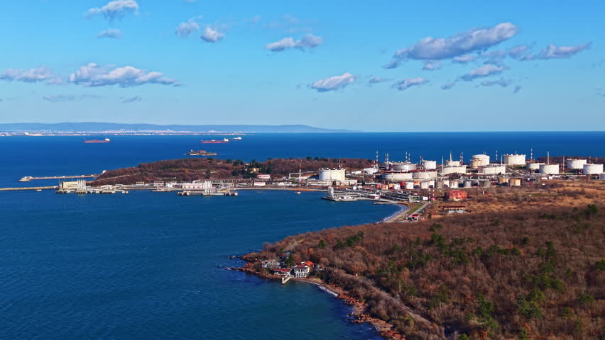 A coastline features an industrial area with storage tanks and a few ships in the water under a bright sky. Trees line the shore, and the sea stretches into the distance.