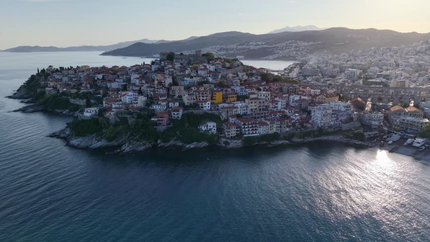 Aerial reveal of historic rooftops, coastal harbor, and reflective bay water in warm evening light.