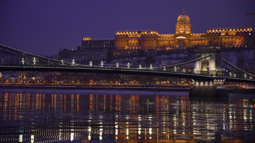 Budapest Night Cityscape with Illuminated Szechenyi Chain Bridge and Buda Castle over the Danube River in Winter, Hungary Budapest, Hungary - January 9, 2026: A wide cinematic tracking shot captures the golden glow of the Szechenyi Chain Bridge and Buda Castle at dusk. The scene features a purple twilight sky, shimmering reflections on the icy Danube River, and a snow-dusted riverbank. This high-quality footage is ideal for travel documentaries, European city marketing, winter tourism promos, and editorial news segments. The composition offers ample copy space in the dark sky.