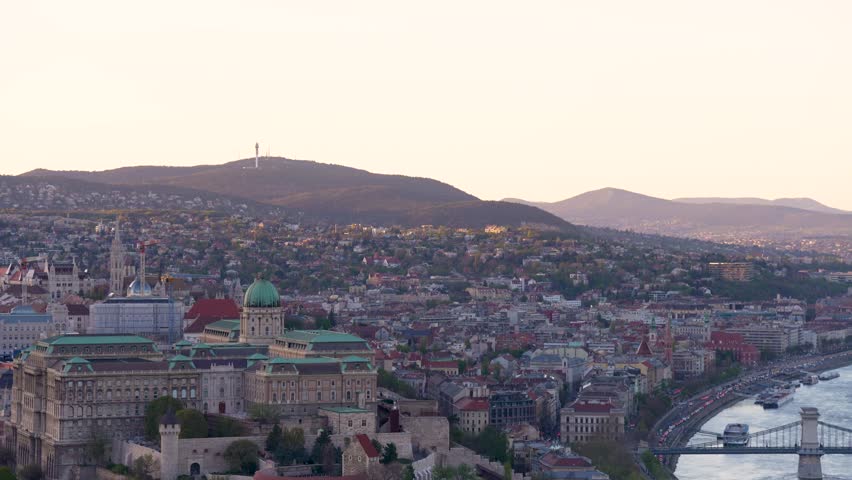 Aerial Panorama of Budapest Hungary at Sunset Featuring Buda Castle Szechenyi Chain Bridge and Danube River with City Skyline and Hills Budapest, Hungary - April 7, 2026: This scenic wide-angle shot pans across the stunning Budapest skyline during the golden hour. The sequence captures the historic Buda Castle and the iconic Szechenyi Chain Bridge spanning the Danube River. Warm, cinematic lighting illuminates the architectural landmarks and the rolling Buda Hills in the background. Ideal for travel documentaries, urban marketing, and editorial features on European heritage.