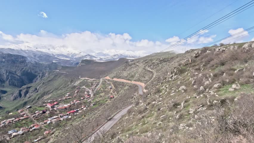 A long mountain road winds through a rural area. Small houses dot the landscape below. Snowy peaks rise in the distance under a bright sky. Nature surrounds the area.