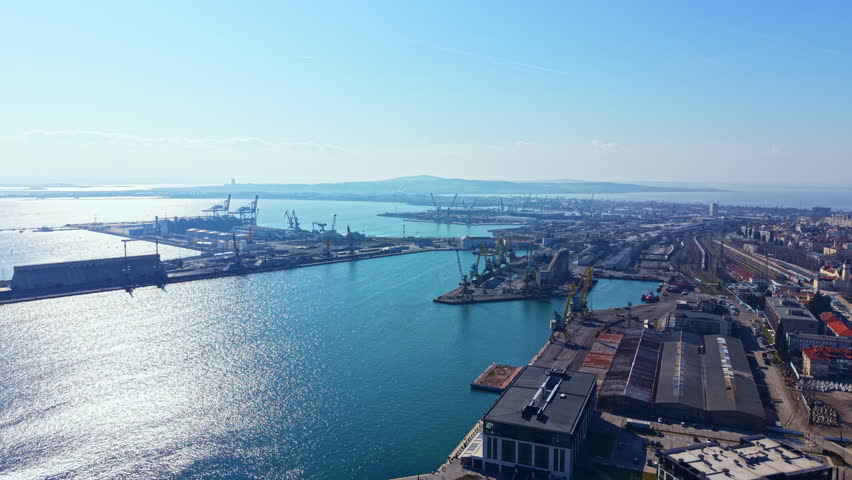 A busy port scene shows ships docked, cranes loading cargo, and industrial buildings near the water. The sun shines bright in the clear sky above the harbor.