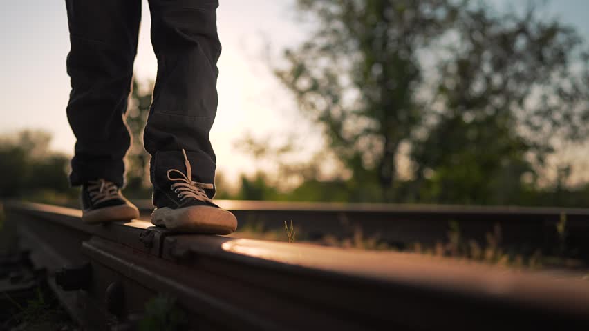 Child is walking on railroad track at sunset. Boy balances on rail in sneakers during summer evening. Legs step on train tracks outdoors. Kid walks away in nature with sun light in background.
