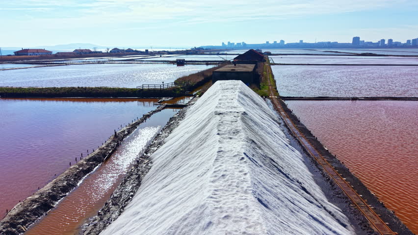Salt mounds are seen on flat areas of water with a view of buildings in the distance during a clear day. The setting shows salt production and landscape.