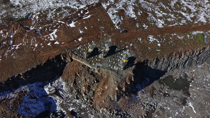 Heavy machinery moves rock and dirt at a granite quarry. Workers focus on tasks while snow covers parts of the ground. The site is busy with activity and machines.