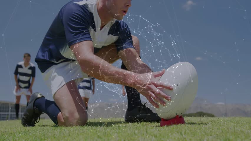 Rugby kicker placing ball on red tee, rising, running up aiming place-kick, particles circling ball. Action, athletic, team, teammates, outdoor, field, grass