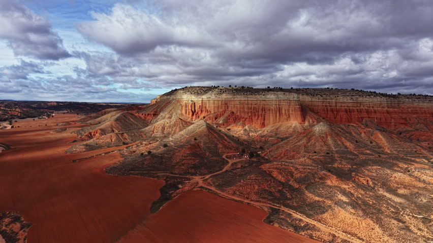 Canyon setting features rocky hills and dry earth with sparse vegetation under a cloudy sky. Shadows fall over the landscape as the sun creates patterns on the ground.