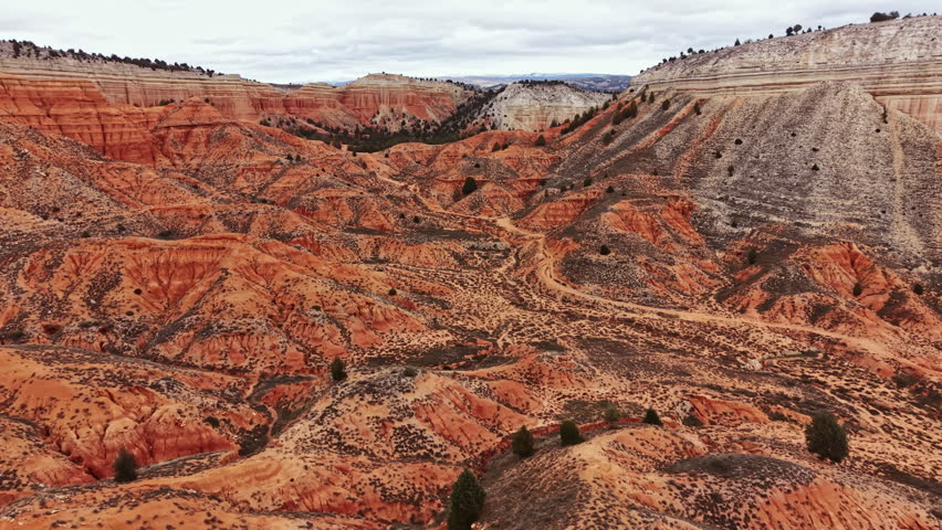 A wide canyon with orange rock formations and paths cutting through the land. The sky is cloudy and the view shows rugged terrain in the distance.