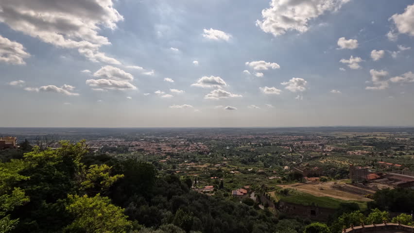 Stunning panoramic view of Tivoli, Italy, overlooking the vast Roman countryside with lush green trees, terracotta rooftops, and dramatic cloud-filled sky stretching toward Rome.