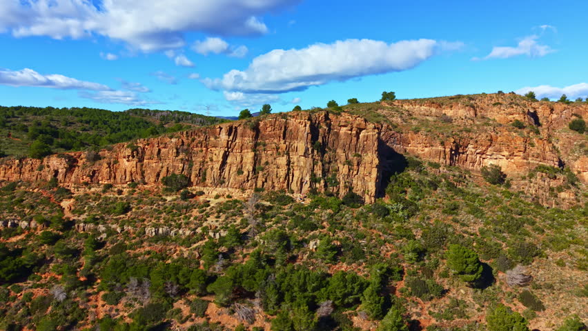 Cliffs rise above a patchy landscape filled with green trees. A bright blue sky with clouds stretches overhead. This scene shows natural beauty in a rural area.