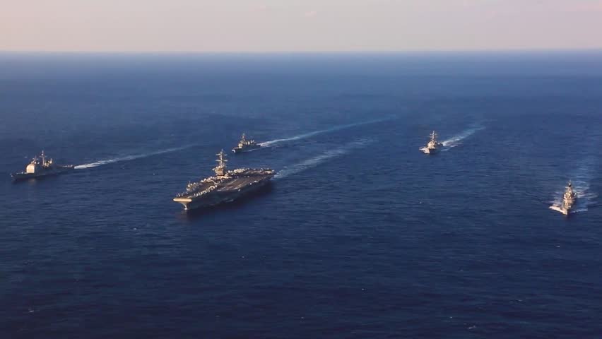 An aerial view of a carrier strike group sailing in formation across the vast blue ocean.