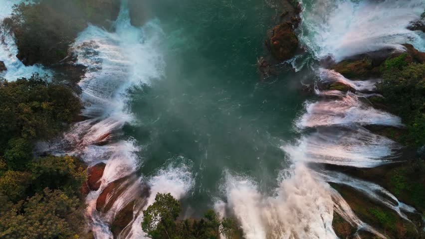 Cinematic drone shot ascending and turning over the majestic Ban Gioc Waterfall. Cascading white water falls into a turquoise river surrounded by lush tropical forest in Northern Vietnam.