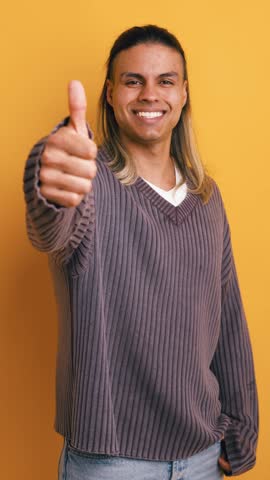 Handsome young man with long dyed hair smiling and looking at the camera. Positive person showing a thumbs up gesture against a yellow background. Vertical