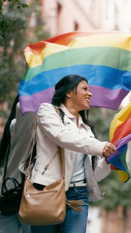 Joyful woman with long dark hair smiling and proudly waving a large rainbow flag during an outdoor lgbt pride parade in the city street. Vertical footage
