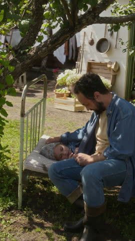 Full vertical shot of loving Caucasian farmer father in rubber boots and denim shirt tenderly kissing little daughter, while putting child for midday sleep on old bed in garden of rural family home