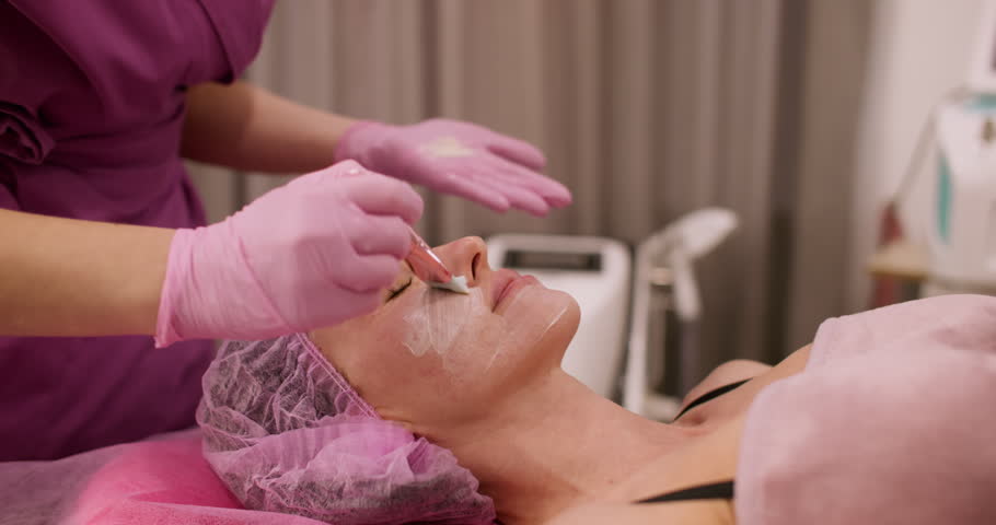 Beauty care. Rejuvenation procedure. Professional female cosmetologist hand using brush applying white clay on woman client face.