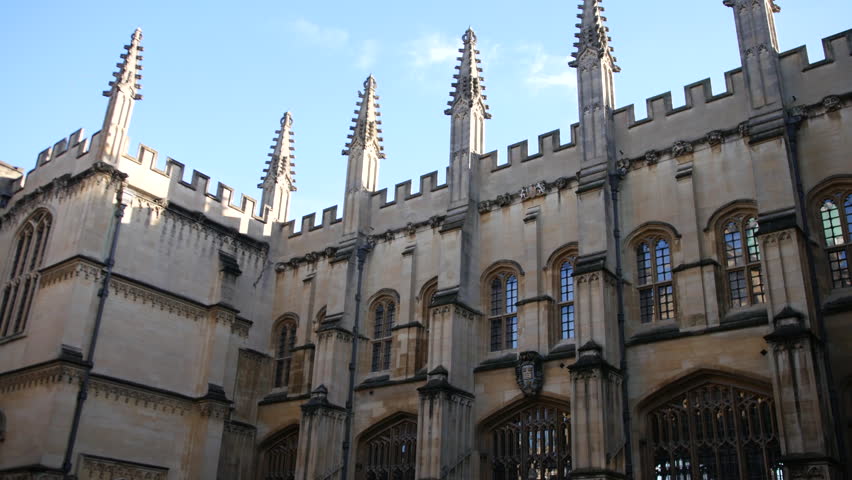 Building courtyard at Oxford University with Gothic spires, arched windows, and stone walls under blue sky, wide angle view highlighting symmetry and traditional college architecture