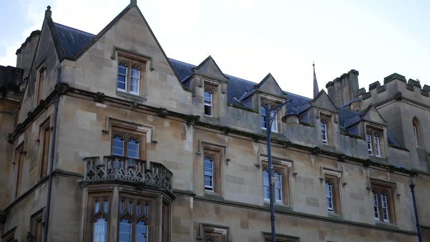 Building at Oxford University with traditional college facade, dormer windows, and stone walls along a city street, classic Oxford architecture captured in wide urban view