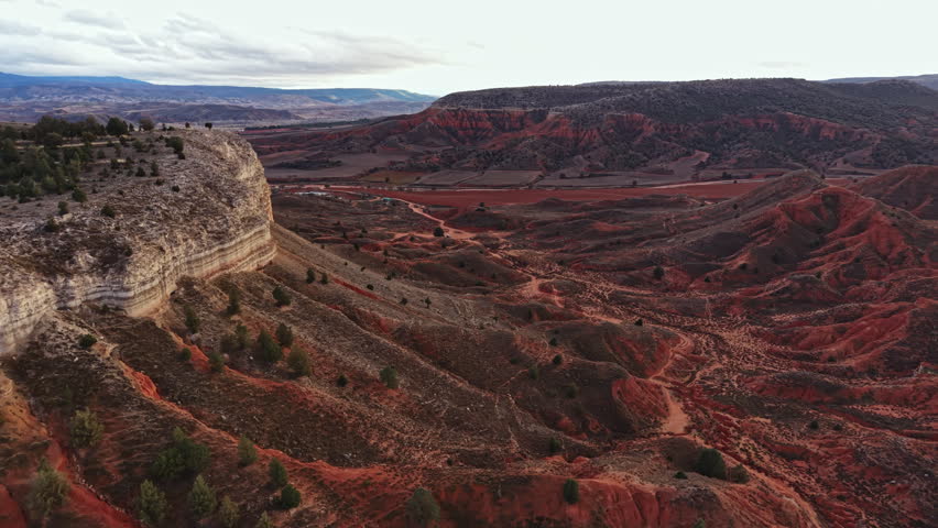 The scene shows a vast area of rocky terrain with red and brown colors. Rolling hills and mountains stretch into the distance under a cloudy sky during late afternoon.