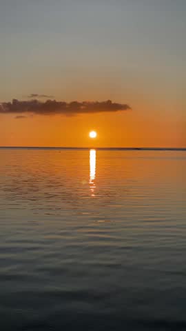 A vertical shot of a vibrant orange sunset over the sea in Mauritius. The sun is low on the horizon casting a bright vertical reflection on the ripples of the water. A single dark cloud floats above the sun against a clear golden sky.