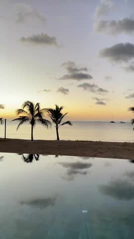 Peaceful tropical scenery in Mauritius at dusk. The calm infinity pool in the foreground reflects clouds and a golden sky. Tall palm trees line a sandy beach facing the Indian Ocean where boats rest on the horizon.
