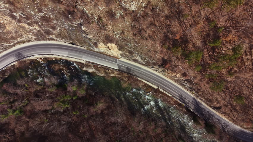 The scene shows a winding road next to a river flowing through a forested area. Trees are visible along the riverbank and rocky cliffs surround the road.