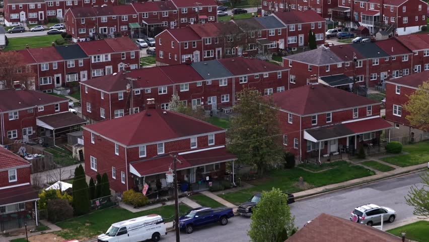 Aerial flyover of brick townhouses and single-family homes in Baltimore suburban neighborhood, showing residential layout, porches and streets during spring in Maryland, USA. Parking cars on roads.