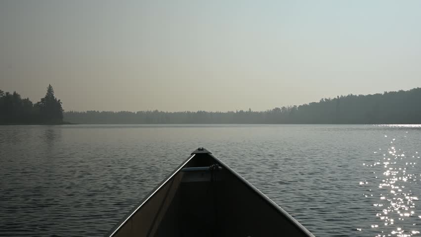 Point of view from a canoe drifting on a smoke covered northern lake with sunlight reflecting across the water in a calm moody wilderness scene.