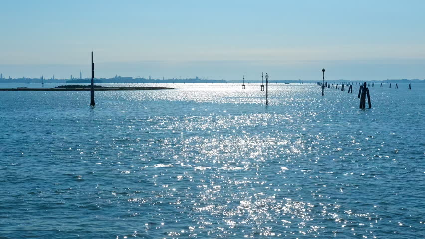 Sunlight reflecting on the venetian lagoon near venice. Bright sunlight reflects on the calm water surface of the venetian lagoon, with navigation poles and the silhouette of venice on the horizon
