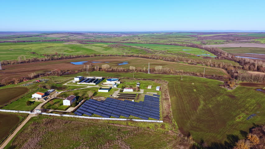 Solar panels cover part of a farm situated in open land. Surrounding fields are green and appear healthy. Utility buildings and water sources are visible nearby under clear weather.