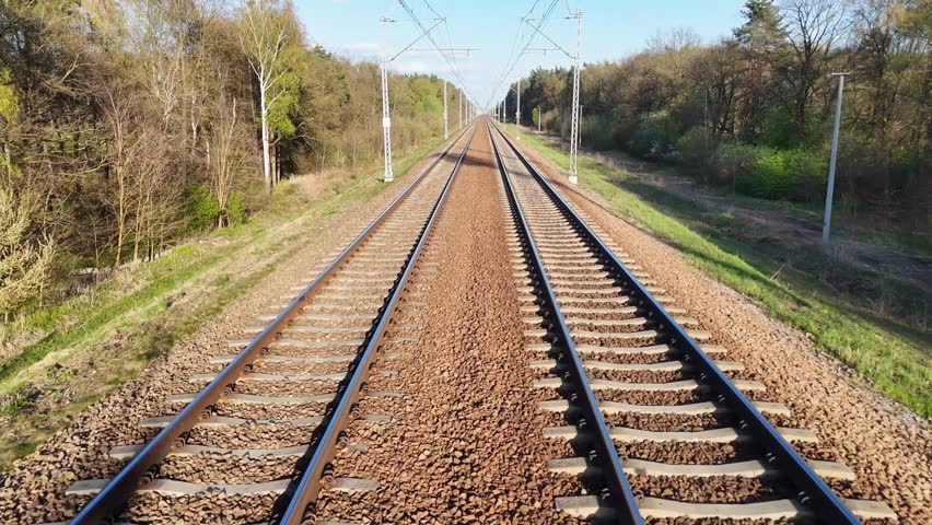 Train tracks stretch into the distance through a lush green landscape, showcasing the gradual transition of scenery along the railway corridor under a clear blue sky