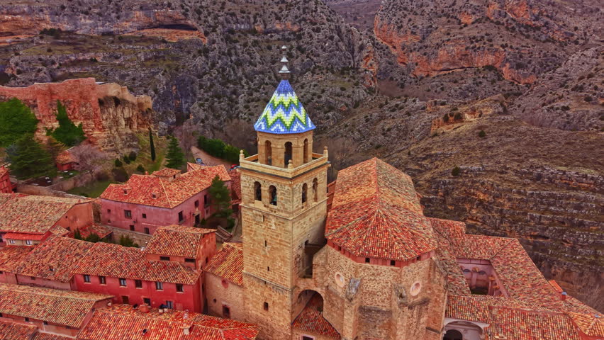 The scene shows an old church with a colorful tower in a rural area with mountains around. The church is part of a small village with stone buildings and red roofs under clear blue sky.