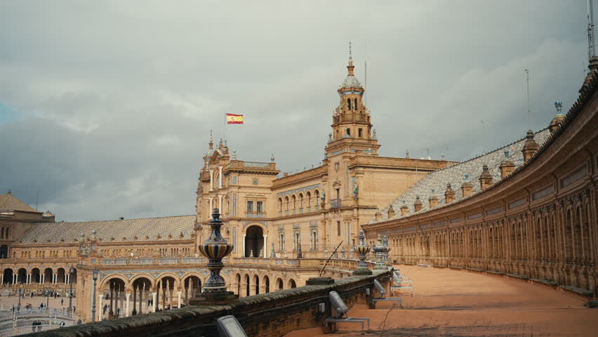 Wide view of Plaza de Espana in Seville, Spain, showcasing its grand semicircular architecture, detailed towers, and Spanish flag under a dramatic cloudy sky.