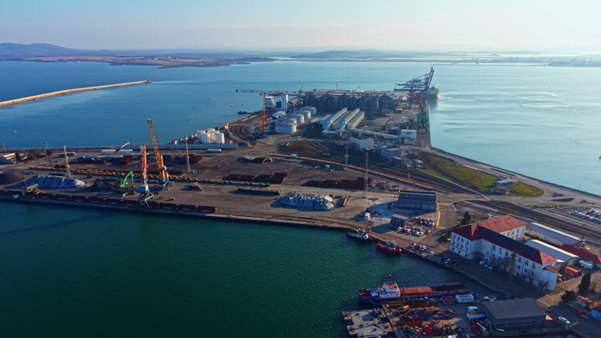 Cargo facilities and ships are seen at a busy port during the day. Cranes and storage areas indicate active loading and unloading of goods by the water.