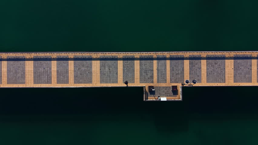 A long wooden pier stretches out over a green lake. People walk along the pier, enjoying the sunny day. The water is clear and calm below the structure.