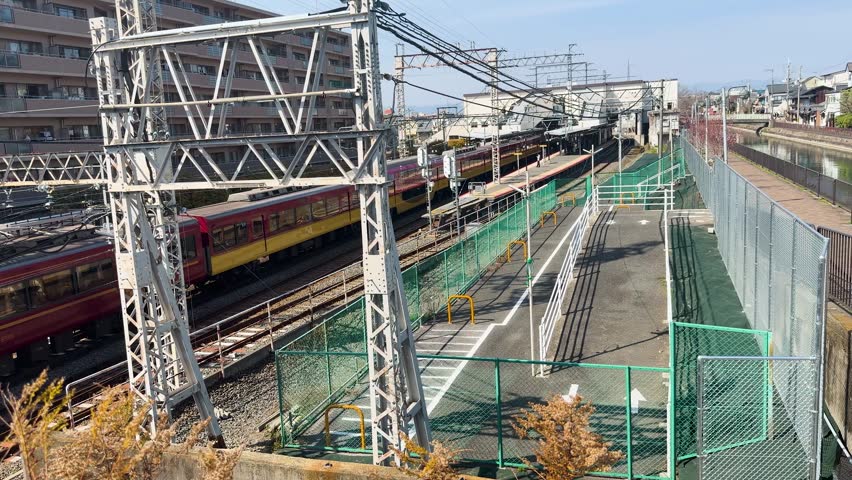 Train passing station, overhead wires, residential buildings, sunny day.
