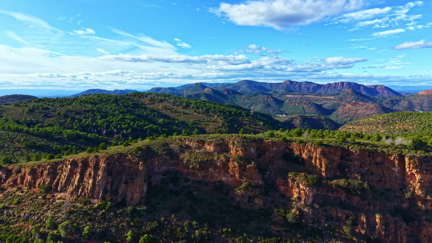 Mountains and cliffs stretch across the landscape, with green trees covering the hills. The sky is bright blue with scattered clouds, capturing nature in daylight.
