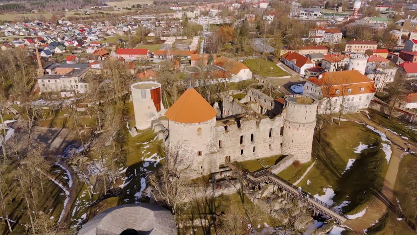 Aerial view of a medieval castle with round towers, stone walls, and bridge approach, surrounded by parkland and town buildings in early spring light.