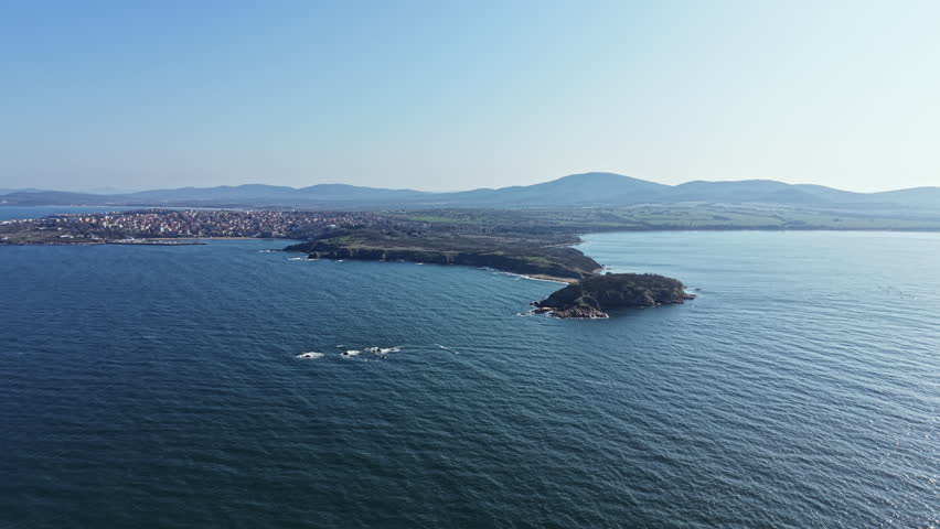 Coastline stretches out towards hills while small waves roll in. A town is visible in the distance. Clear skies reveal a sunny day. The landscape shows harmony of land and sea.