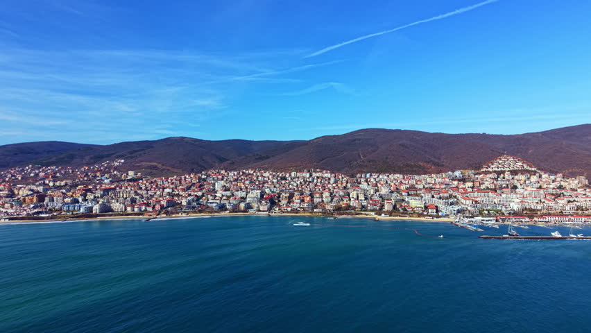 Houses sit on the hills above a beach, with boats in the water below. The sky is clear and blue. This scene shows the lively atmosphere of the coastal town during afternoon.
