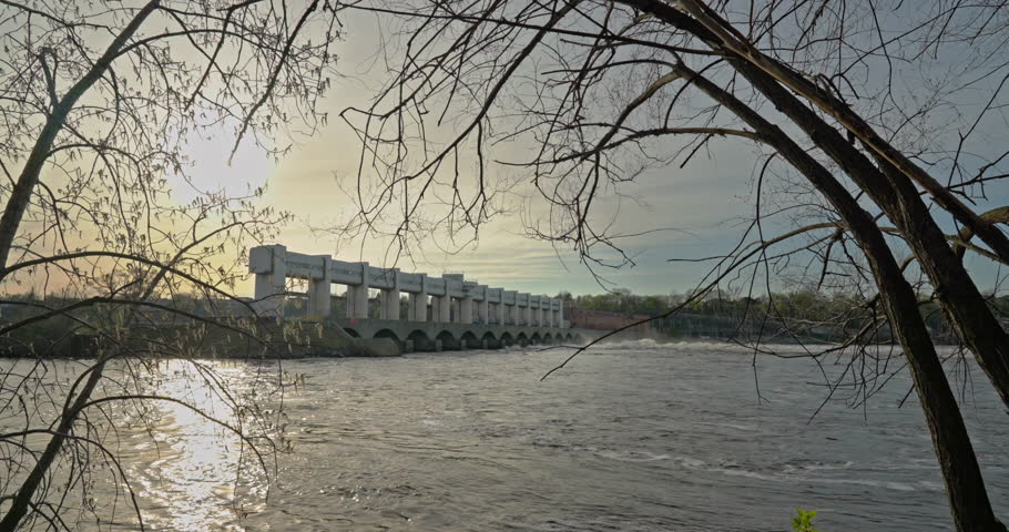 A hydroelectric dam spans a swollen river as strong current sweeps through an early spring high-water scene framed by bare branches and bright seasonal light.
