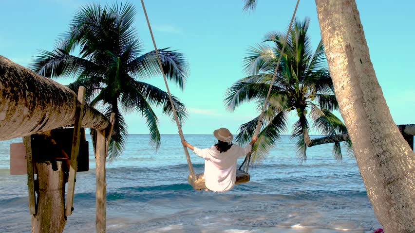 A serene moment captures a woman on a swing on Koh Kood Island, surrounded by lush palm trees and the gentle waves of the sea. The tranquil scene invites relaxation and escape in Thailand