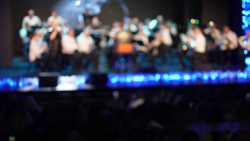 Blurred view of a jazz orchestra performing on a stage with blue lights. Audience in a concert hall enjoying live music in a cozy atmosphere