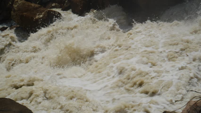 Raging river water crashes over rocks, creating white froth. Turbulent flow shows powerful natural force. Ideal for adventure, nature, or extreme weather themes. No people present