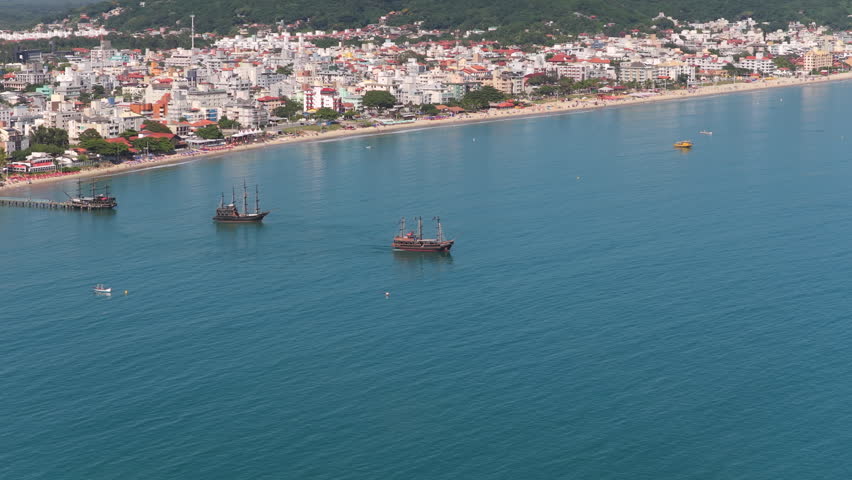 Pirate style tour boats departing Canasvieiras in Florianópolis Santa Catarina Brazil navigating Baía Norte for coastal sightseeing experiences with urban shoreline backdrop, drone orbiting